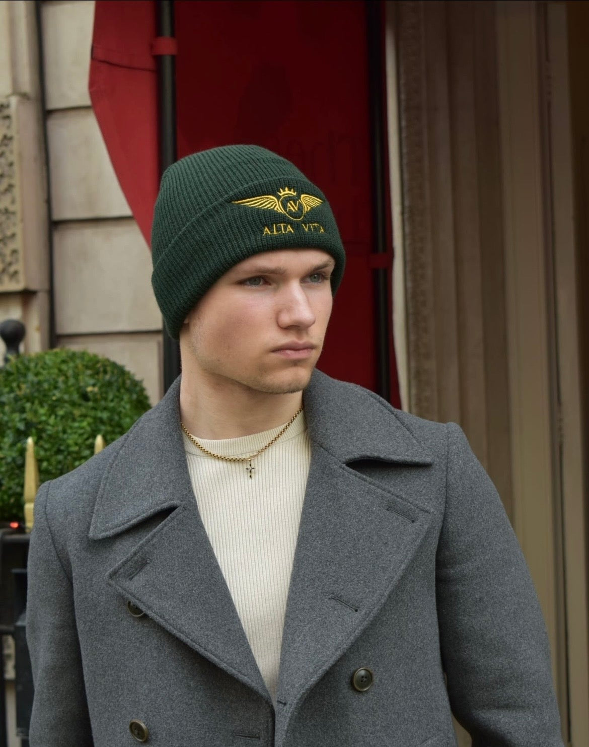 A man wearing a dark green knit beanie with a gold military-style insignia and the text 'Alta Vita' embroidered on the front, paired with a gray double-breasted coat over a cream sweater.